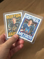A hand holds two Culyer Smith trading cards in plastic sleeves, featuring illustrated players. One card says BOSTON RAIDERS, the other says MONTREAL METROS—both part of the sought-after Heated Rivalry set. Each player has a captain’s C on their jersey, and these cards are a signed numbered edition.