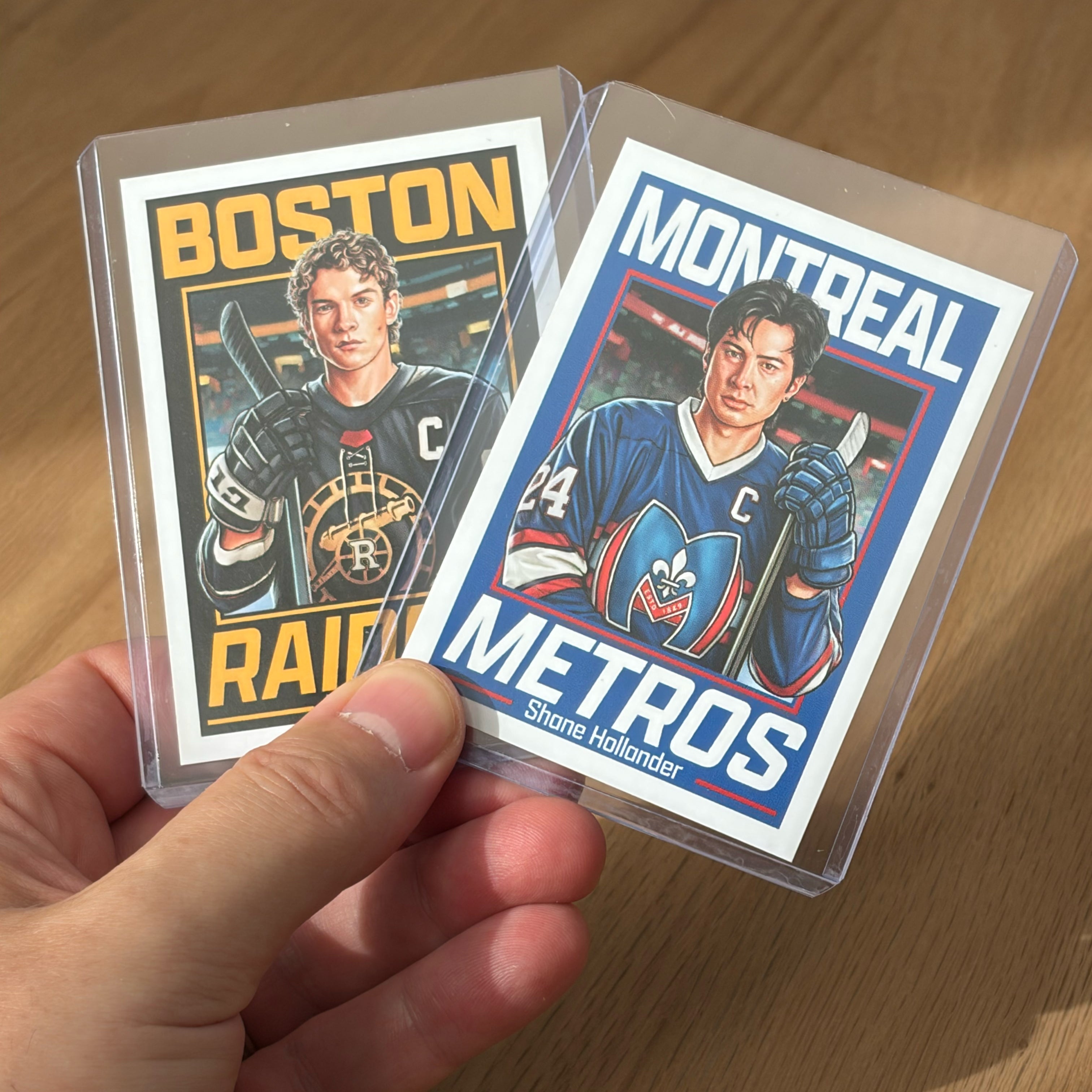 A hand holds two Culyer Smith trading cards in plastic sleeves, featuring illustrated players. One card says BOSTON RAIDERS, the other says MONTREAL METROS—both part of the sought-after Heated Rivalry set. Each player has a captain’s C on their jersey, and these cards are a signed numbered edition.