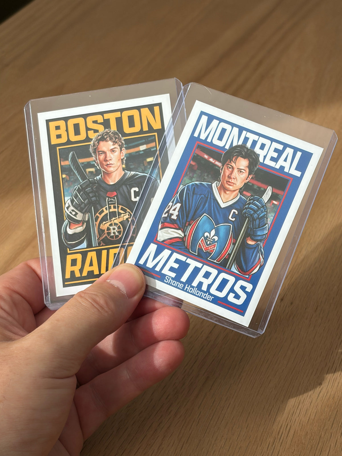 A hand holds two Culyer Smith trading cards in plastic sleeves, featuring illustrated players. One card says BOSTON RAIDERS, the other says MONTREAL METROS—both part of the sought-after Heated Rivalry set. Each player has a captain’s C on their jersey, and these cards are a signed numbered edition.