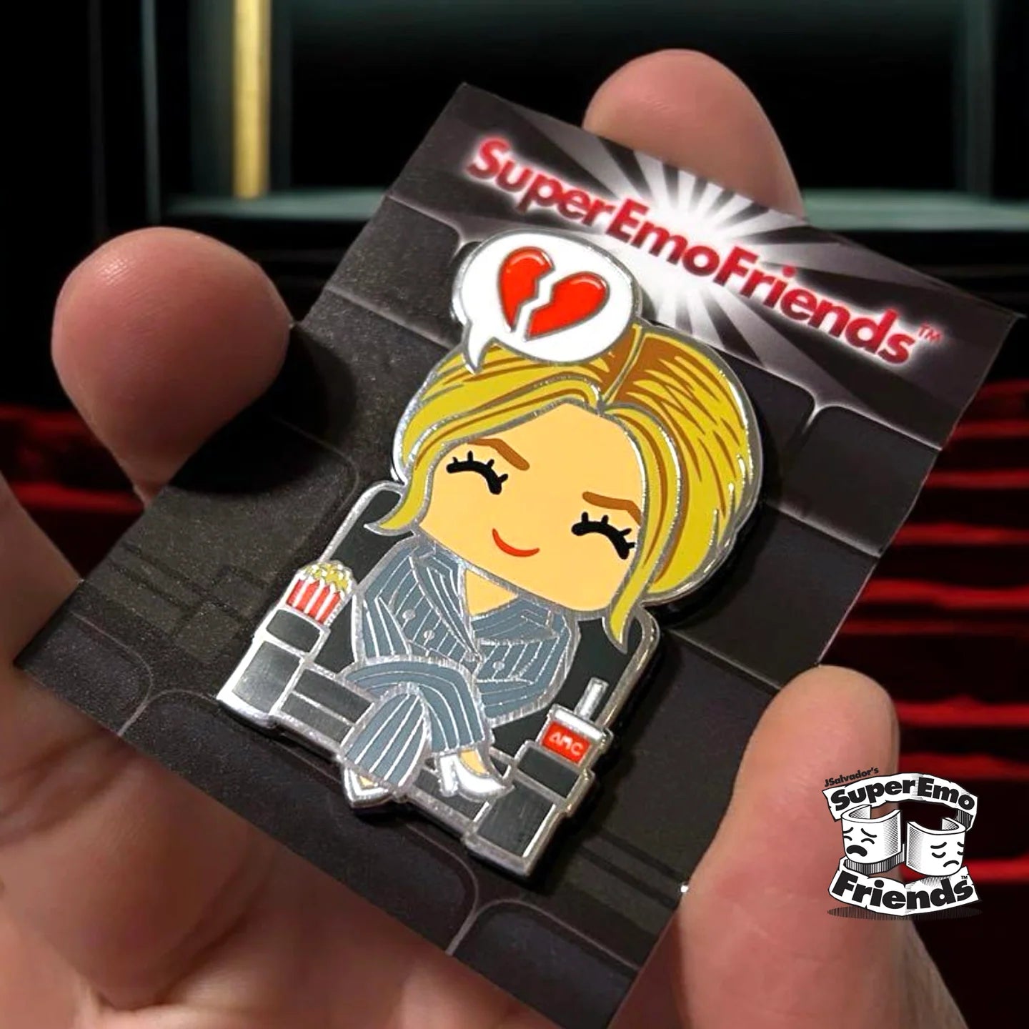 A hand holds a Super Emo Friends limited edition hard enamel pin showing a cartoon character with blonde hair in a striped suit, sitting in a chair holding a drink, beneath a broken heart and the “SuperEmoFriends” text.