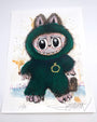 A whimsical Joey Feldman illustration of a furry, wide-eyed creature in a green cat-eared costume with a gold ring necklace, set on a white background with ink splatters, colorful accents, and hand-painted details.