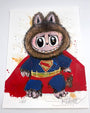 Joey Feldman-inspired illustration of a cute, wide-eyed creature in a Superman costume with a furry hood and bear ears, seated on a white background with colorful paint splatters and hand-painted details.