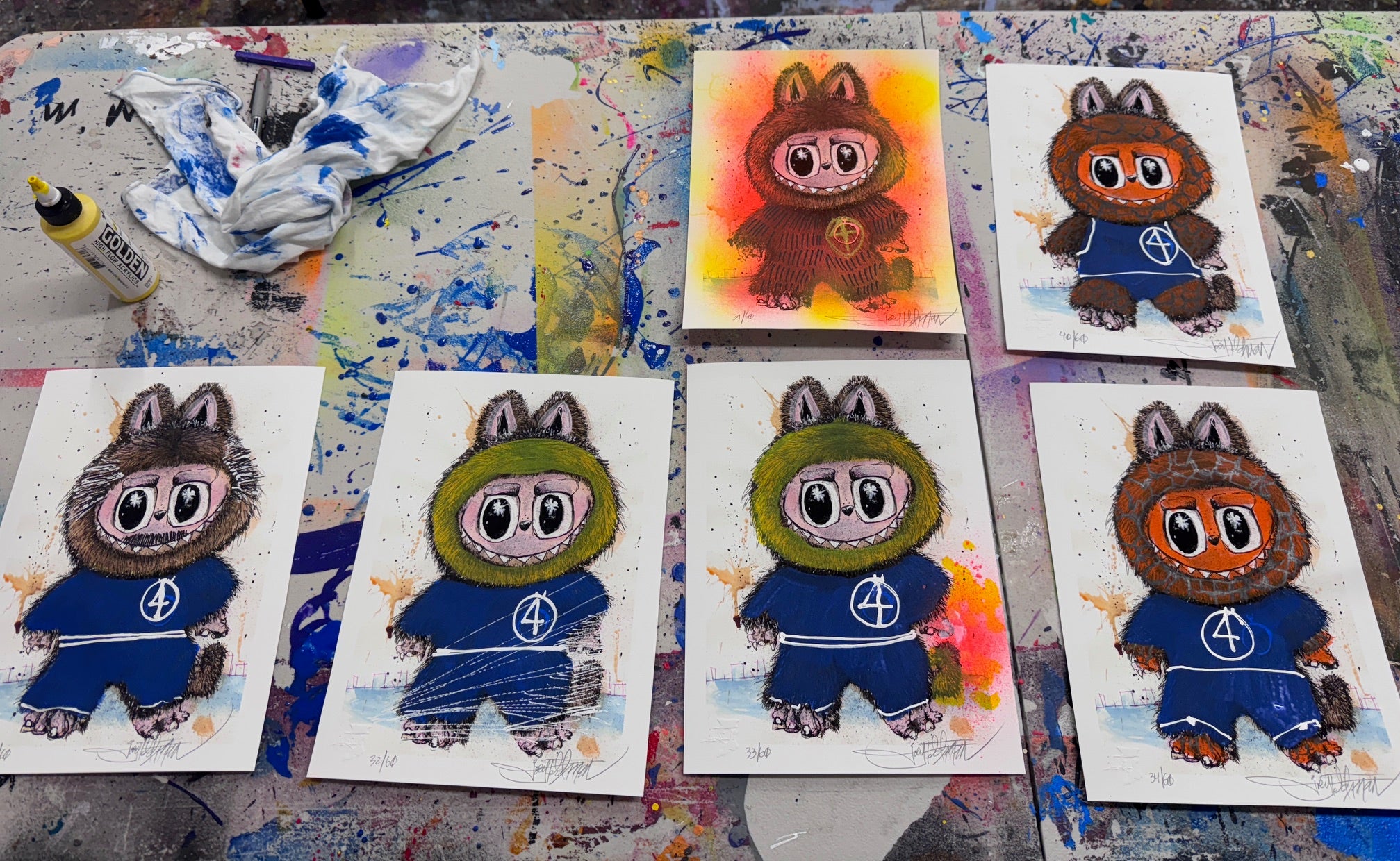 Seven Joey Feldman illustrated cards of a cartoon creature in a blue Fantastic Four sweater are displayed on a colorful, paint-splattered surface. Each archival pigment print features unique backgrounds and hand-painted details. A bottle and cloth sit nearby.