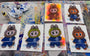 Seven Joey Feldman illustrated cards of a cartoon creature in a blue Fantastic Four sweater are displayed on a colorful, paint-splattered surface. Each archival pigment print features unique backgrounds and hand-painted details. A bottle and cloth sit nearby.