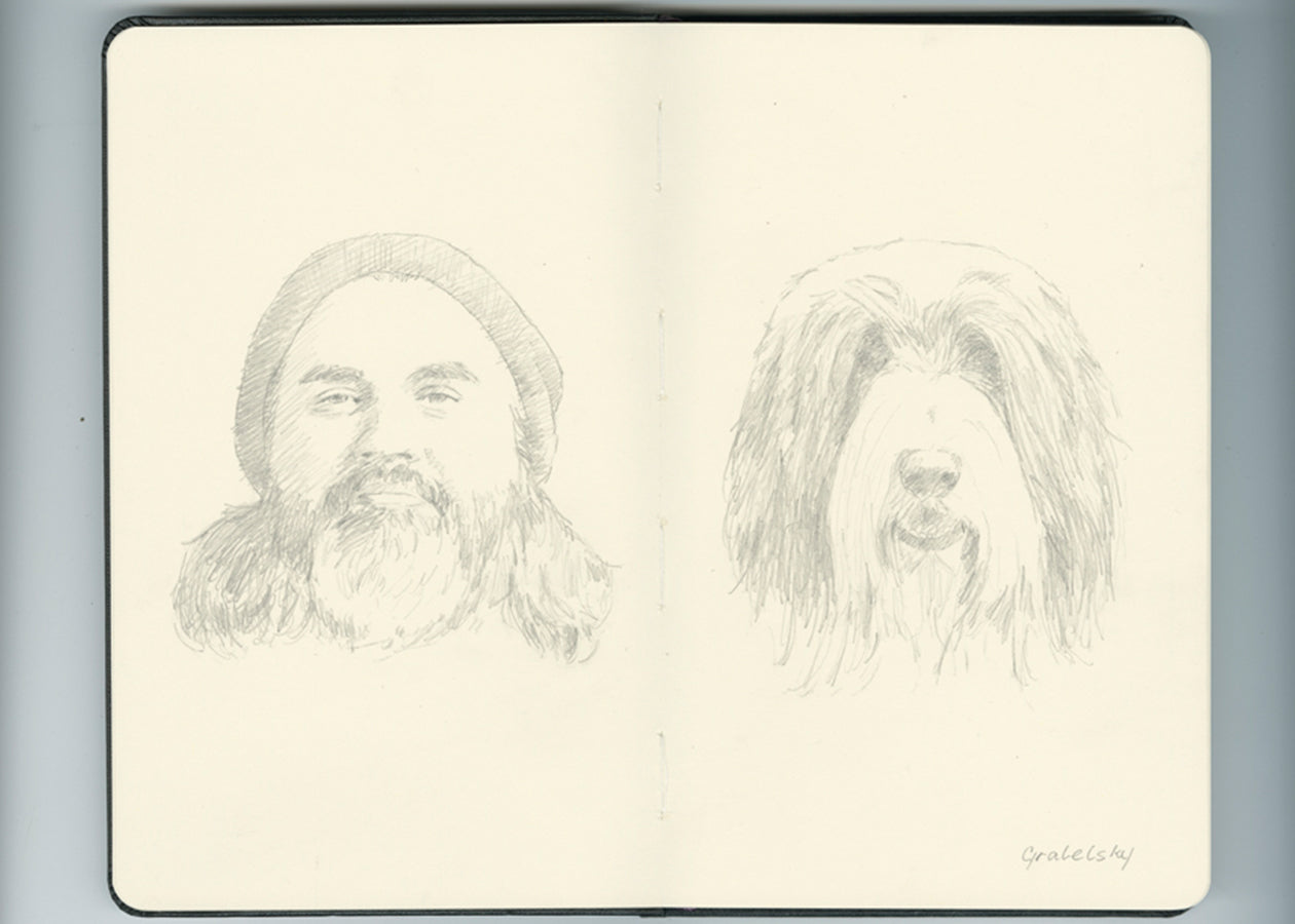 pencil sketch in an open face Moleskine notebook of a heavily bearded man with long hair wearing a beanie on one side, and on the other side a shaggy long-haired dog by artist Matthew Grabelsky