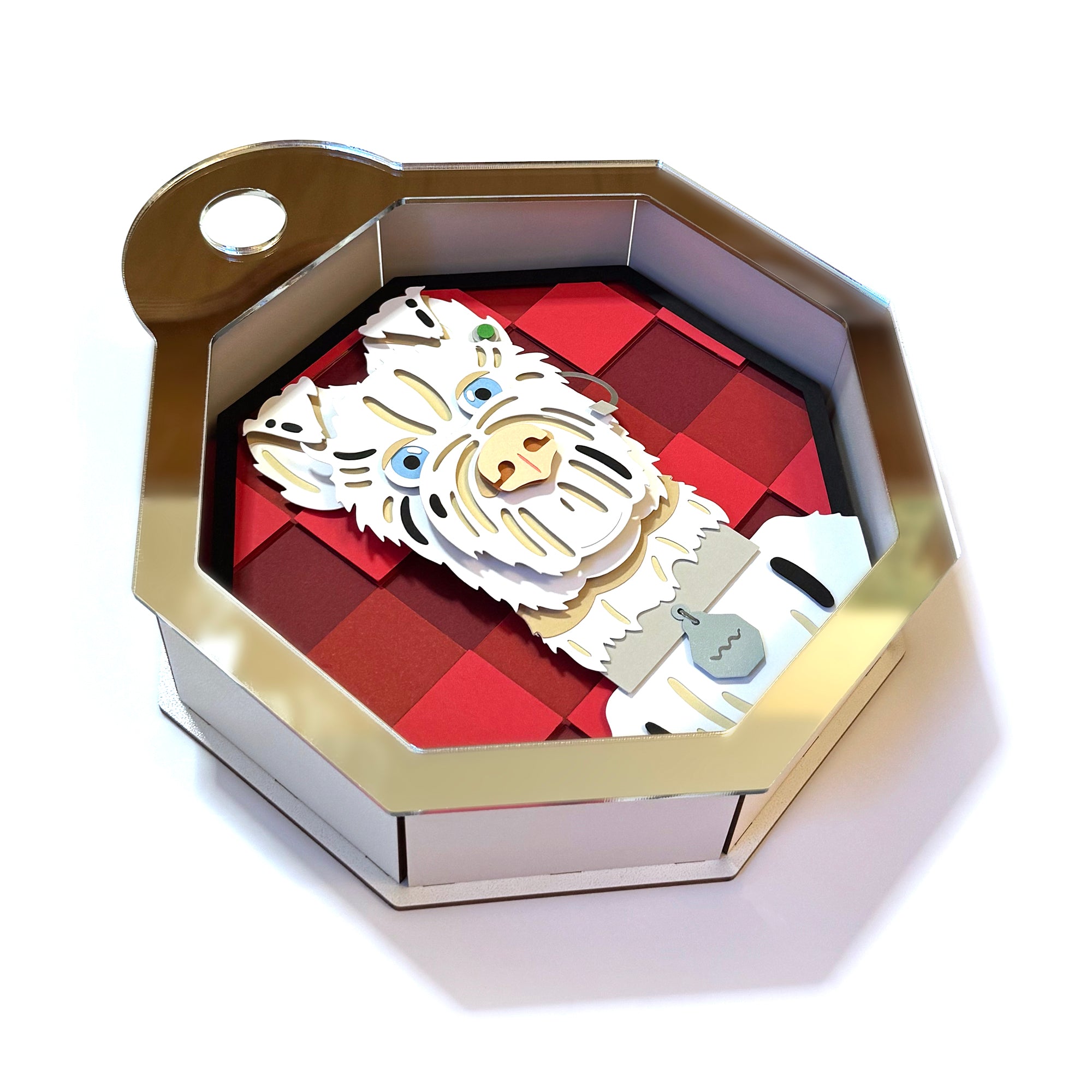An octagonal metallic box by Michelle Romo displays layered paper art of an alpacas head against a red and black checkered backdrop. The alpaca wears a gray scarf, echoing whimsical Isle of Dogs styles, and the box includes a convenient top loop.