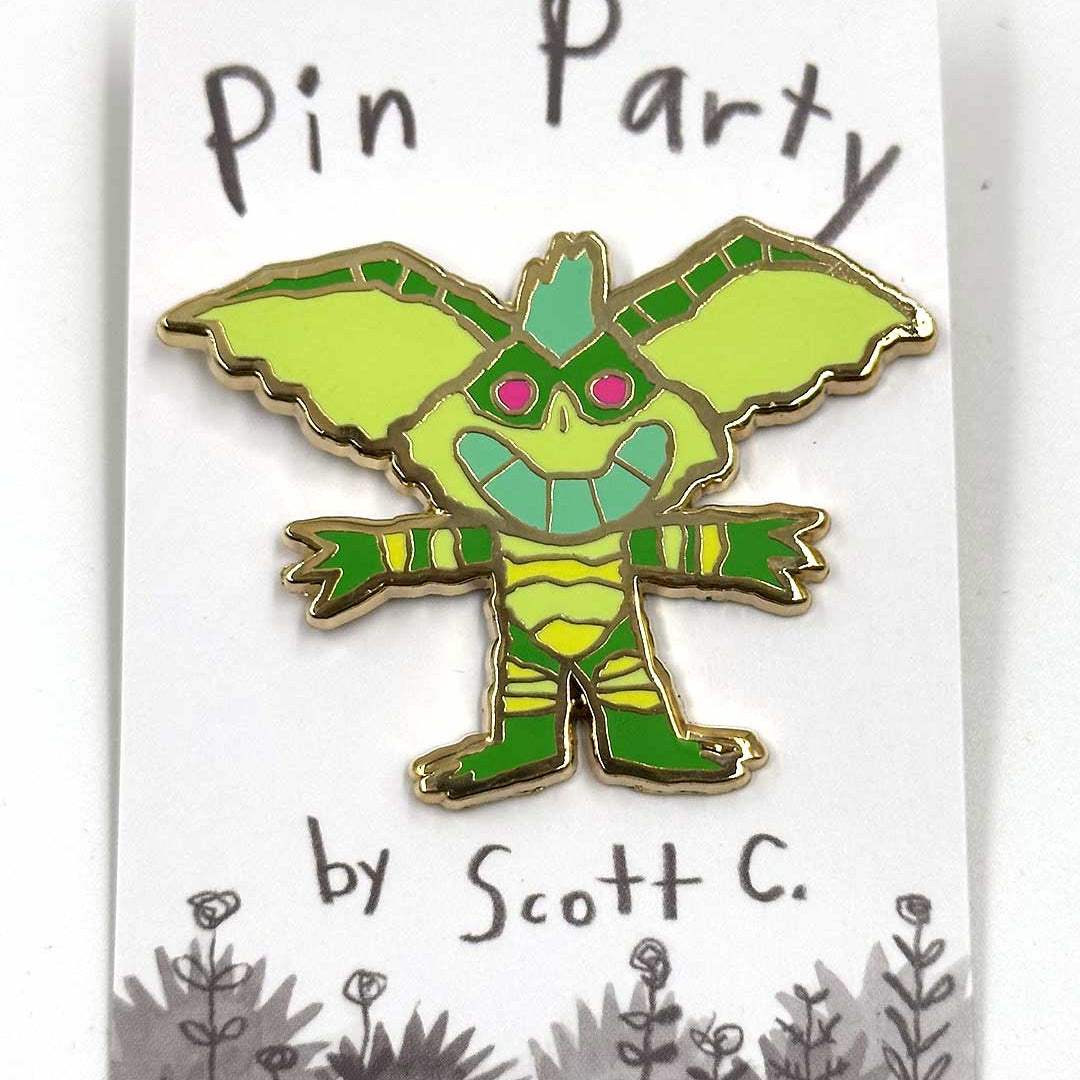 A green, cartoon-style gremlin hard enamel pin with pink eyes is displayed on a card that features the Scott C. brand name and playful flower doodles along the bottom, accentuating this delightful gold metal plated accessory.