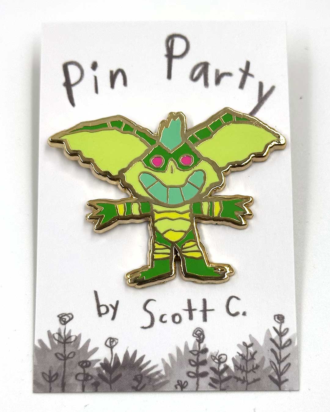 A green, cartoon-style gremlin hard enamel pin with pink eyes is displayed on a card that features the Scott C. brand name and playful flower doodles along the bottom, accentuating this delightful gold metal plated accessory.
