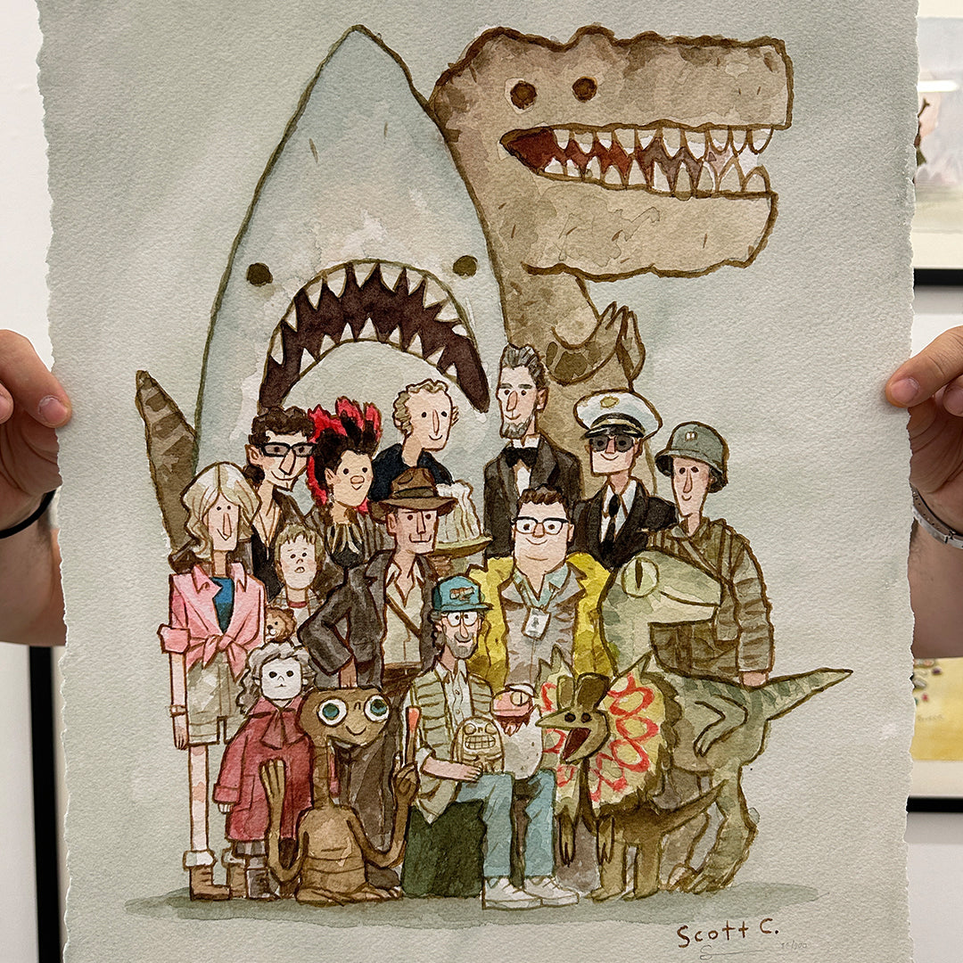 A person holds a limited edition archival pigment print by Scott C. featuring cartoon versions of iconic movie characters—shark, dinosaurs, aliens, and humans—posing for a group photo. Signed by the artist.
