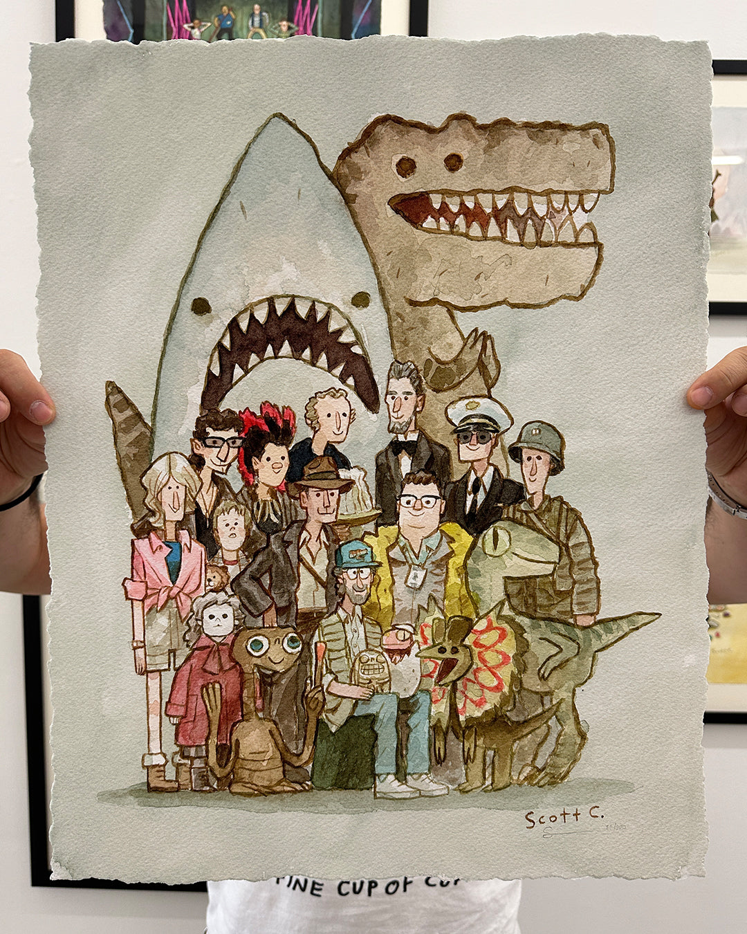 A person holds a limited edition archival pigment print by Scott C. featuring cartoon versions of iconic movie characters—shark, dinosaurs, aliens, and humans—posing for a group photo. Signed by the artist.