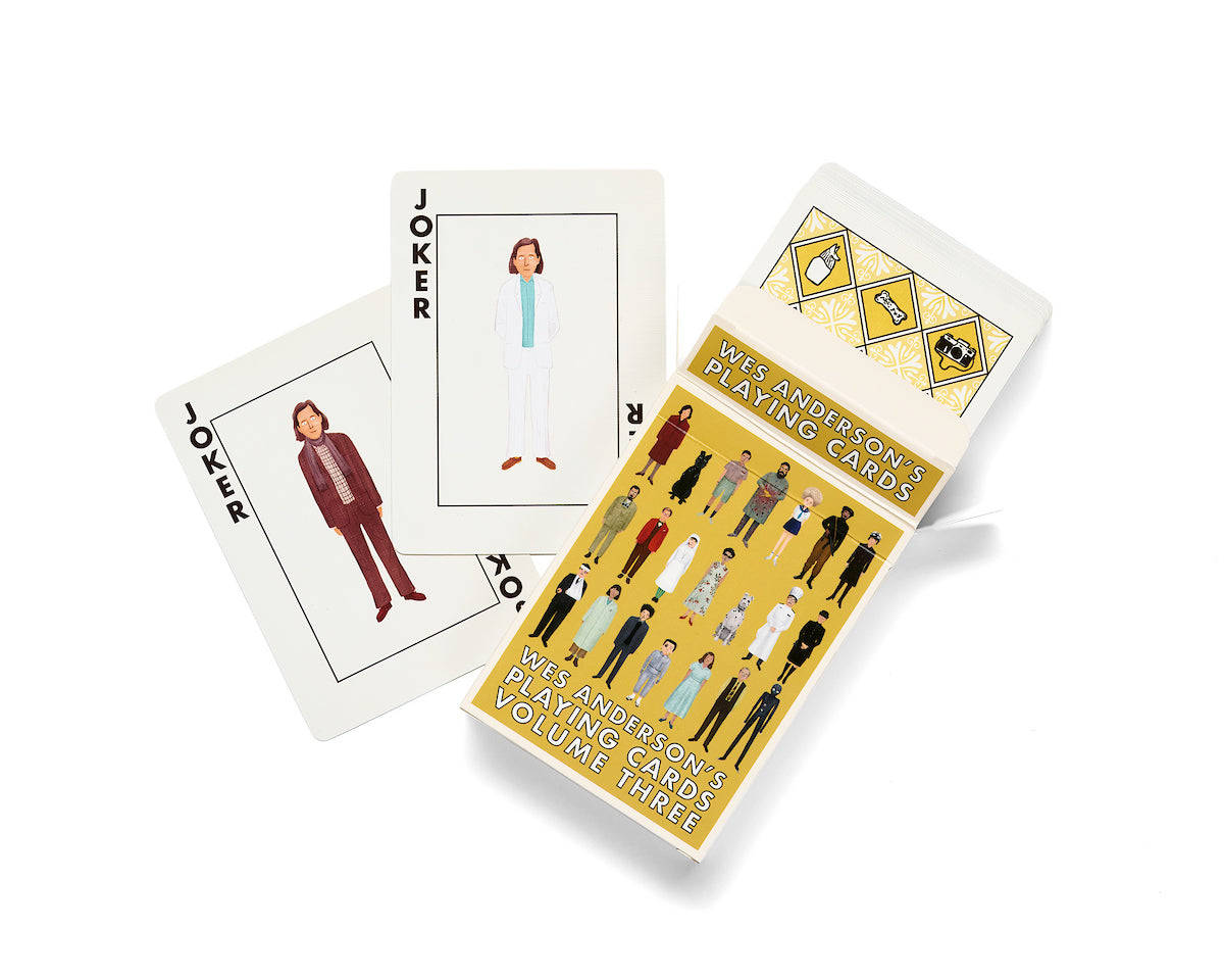Two illustrated playing cards marked “Joker” depict people in suits. An open box labeled "Max Dalton Wes Anderson Playing Cards Volume Three" displays more custom-designed playing cards by Max Dalton, featuring illustrated characters set against a yellow background.