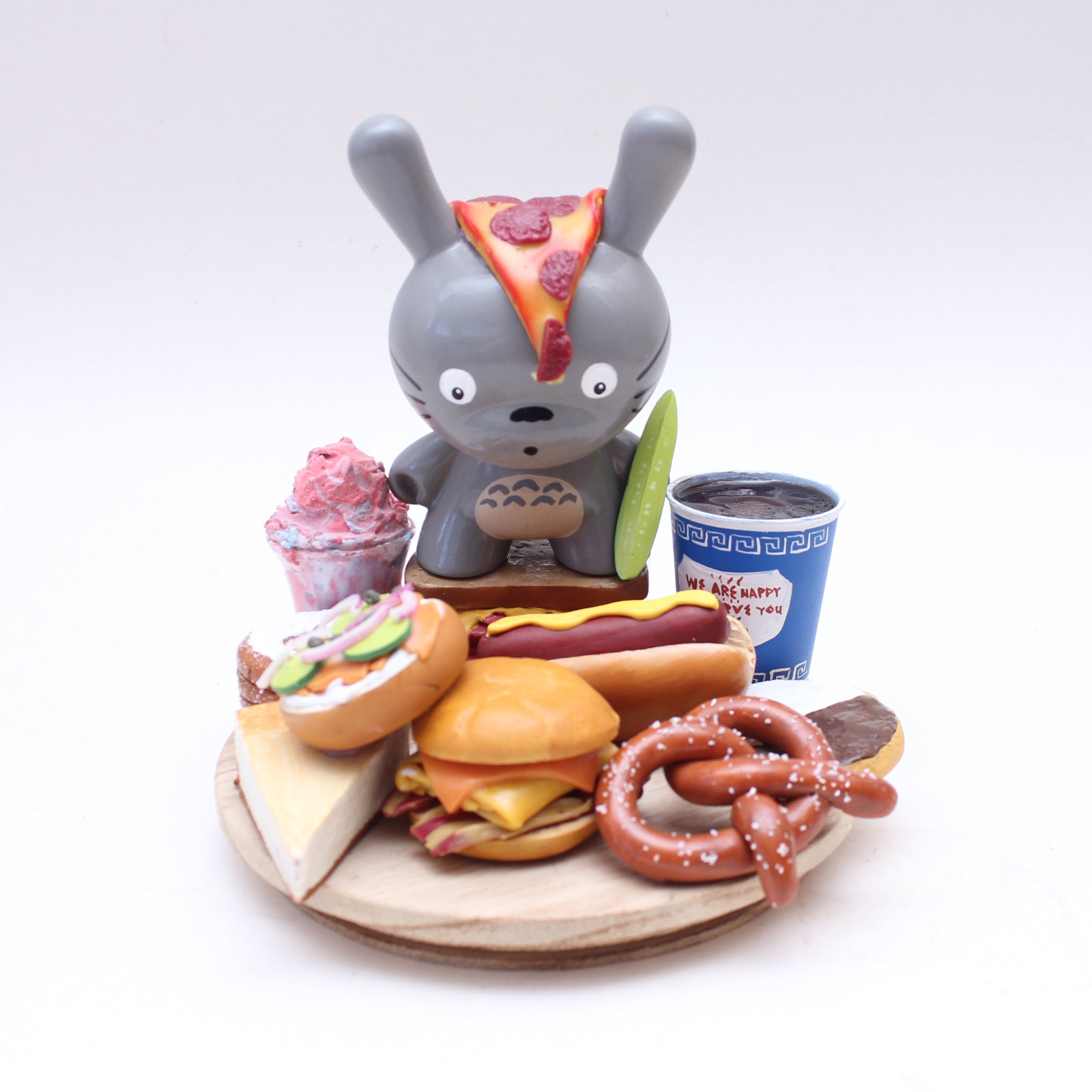 A whimsical mixed media art piece by Spoke Art: a gray bunny toy with a pizza-slice ear stands on a wooden plate, surrounded by toy foods—pretzel, hot dog, burger, sandwich, cheesecake, pink ice cream, and a blue takeout cup.