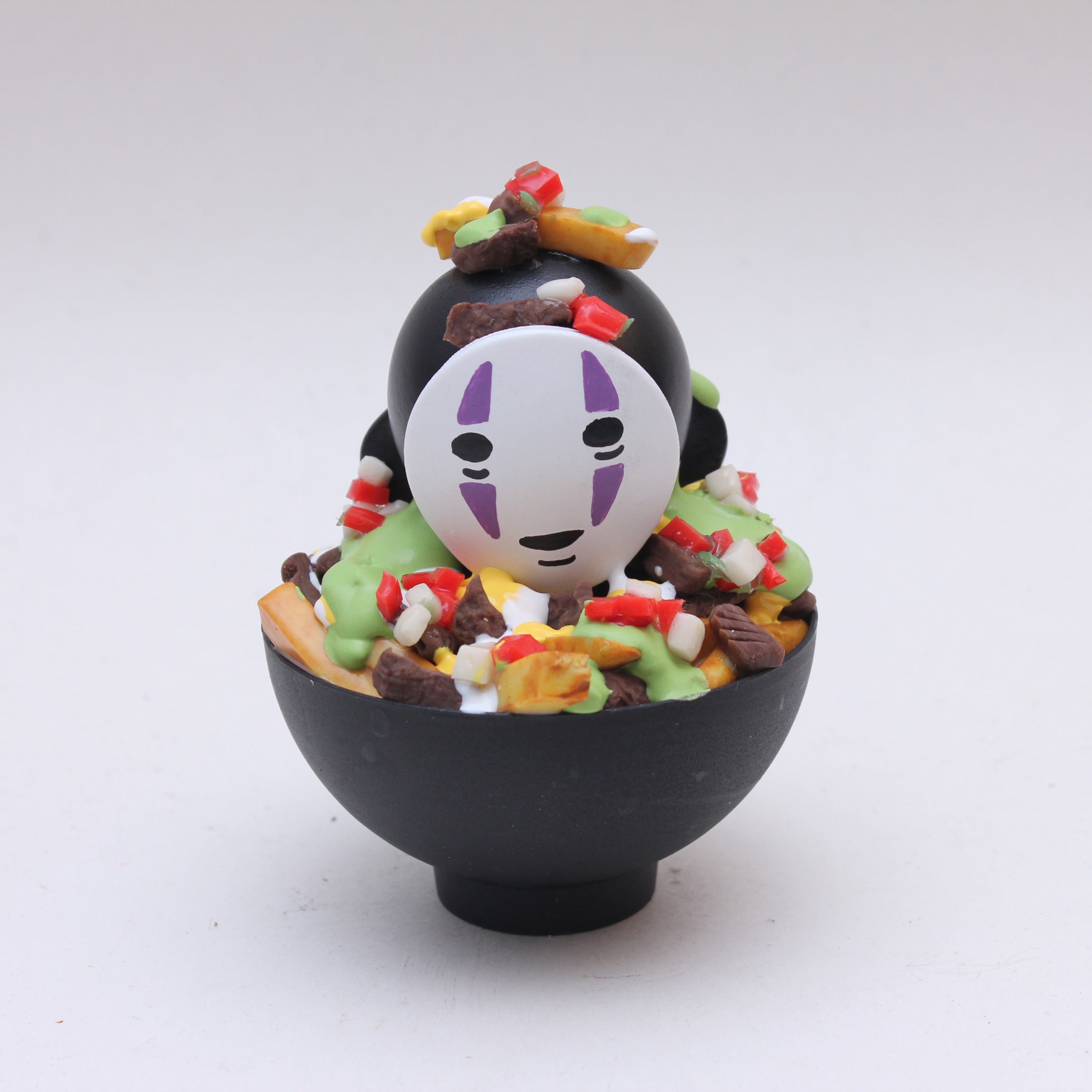 A black bowl by Zard Apuya holds a No-Face-inspired figurine, resembling a Kidrobot Munny vinyl toy, topped with colorful accents to create playful No Face Carne Asada Fries—a unique fusion designed for San Diego Comic-Con 2025.