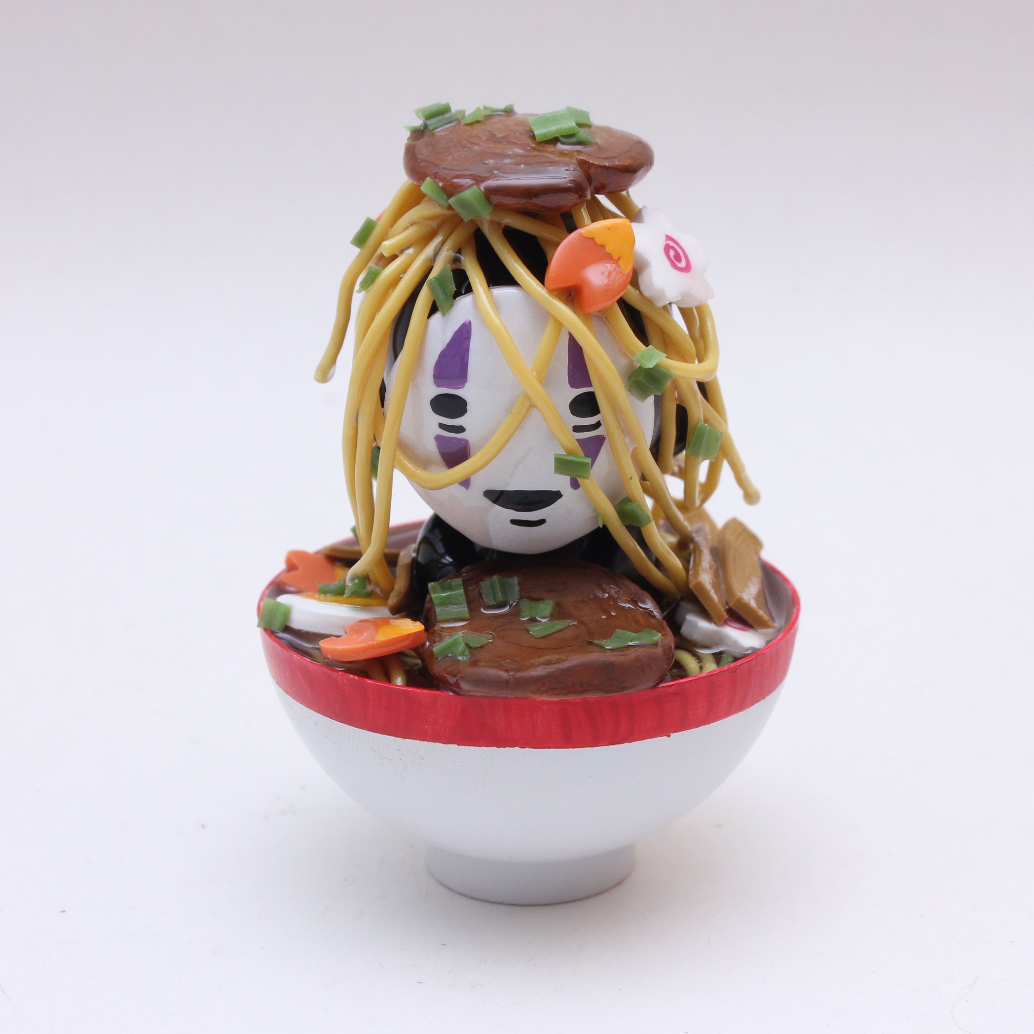 A small figurine with purple facial markings, topped with noodles, vegetables, and meat, sits in a white bowl with a red rim—Zard Apuya’s ramen-inspired vinyl toy featured at San Diego Comic-Con 2025.