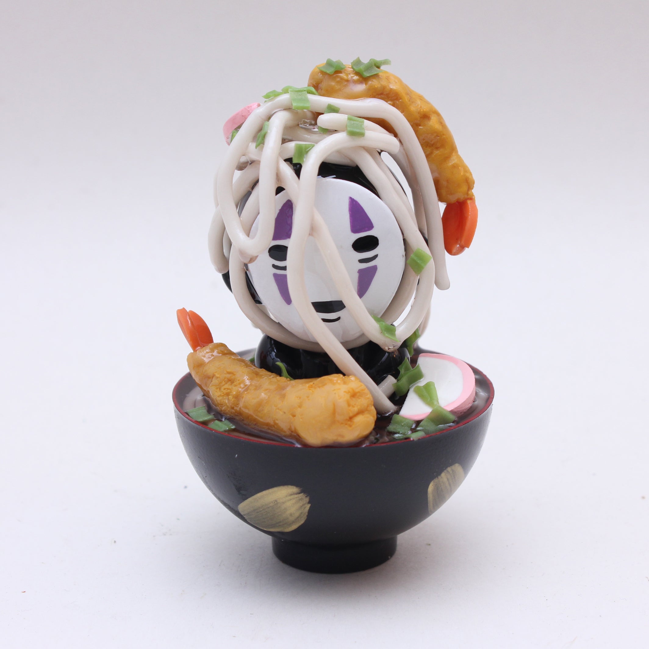 A small Zard Apuya figurine of No-Face sits in a black bowl with shrimp tempura, noodles, and green onions—capturing the charm of a No-Face Udon Bath. Perfect for anime lovers and Kidrobot Munny collectors.