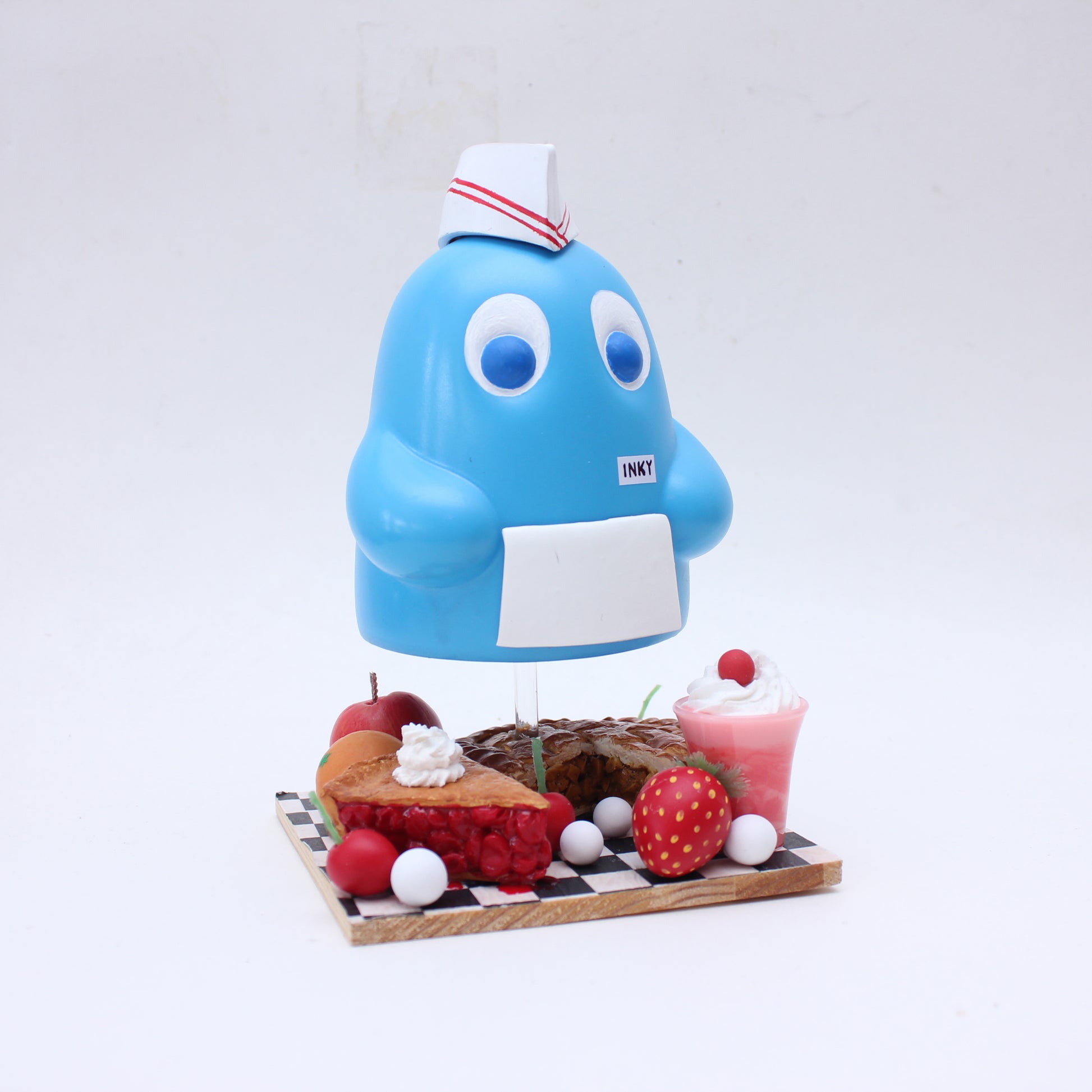 A playful blue vinyl toy by Zard Apuya, featuring big eyes, a white apron, and chef hat, stands on a checkered board with desserts and fruit—perfect for any video game-inspired exhibition.