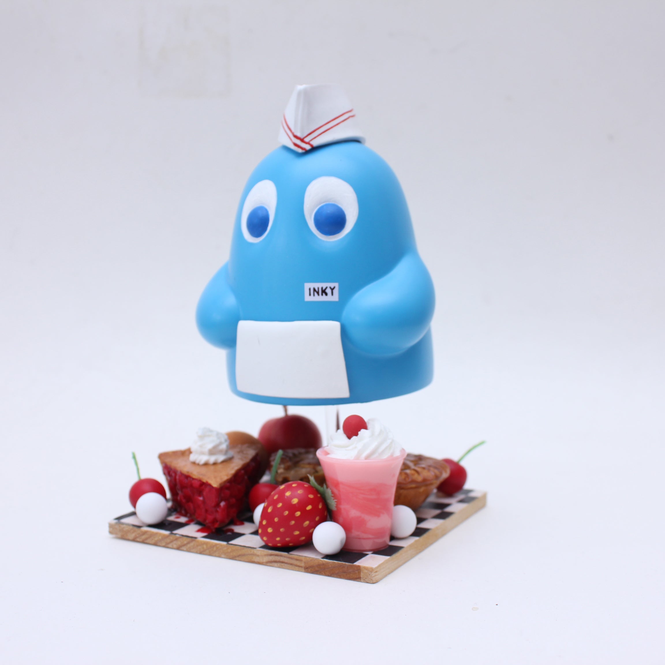 A playful scene features a blue cartoon character with a white hat and an "INKY" tag by Zard Apuya, standing on a checkered platform among desserts, inspired by Pac Man video game art.