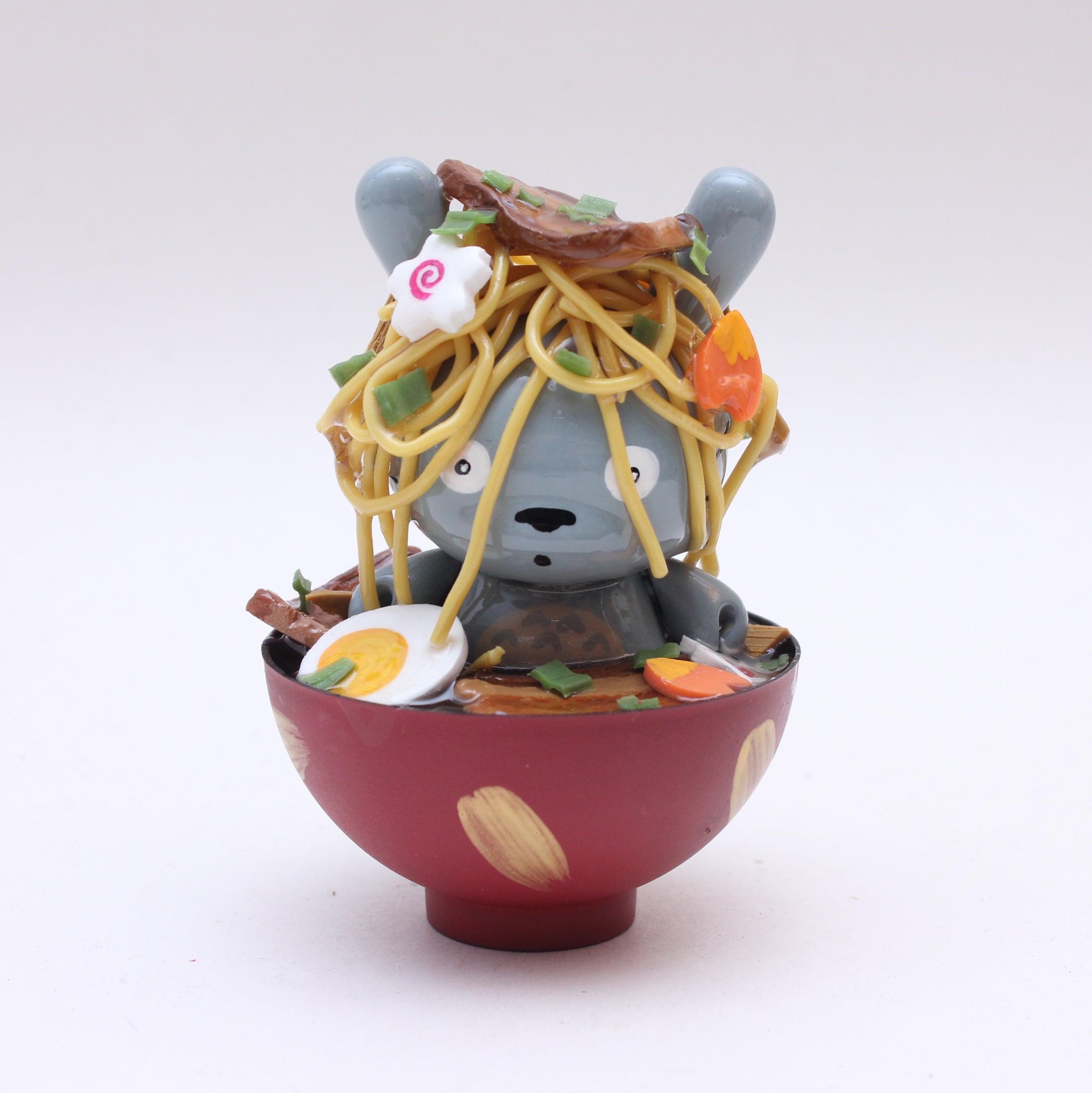 A Zard Apuya gray bear figurine, inspired by Kidrobot Munny toys, sits in a red ramen bowl filled with toy noodles, veggies, and egg, looking surprised against a plain white background.