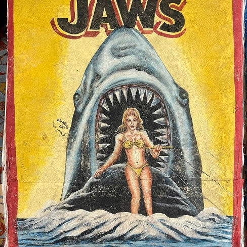 A vintage-style JAWS painting by Deadly Prey depicts a woman in a yellow bikini standing in water as a giant shark with an open mouth looms behind her; the background features bold yellow and red hues.