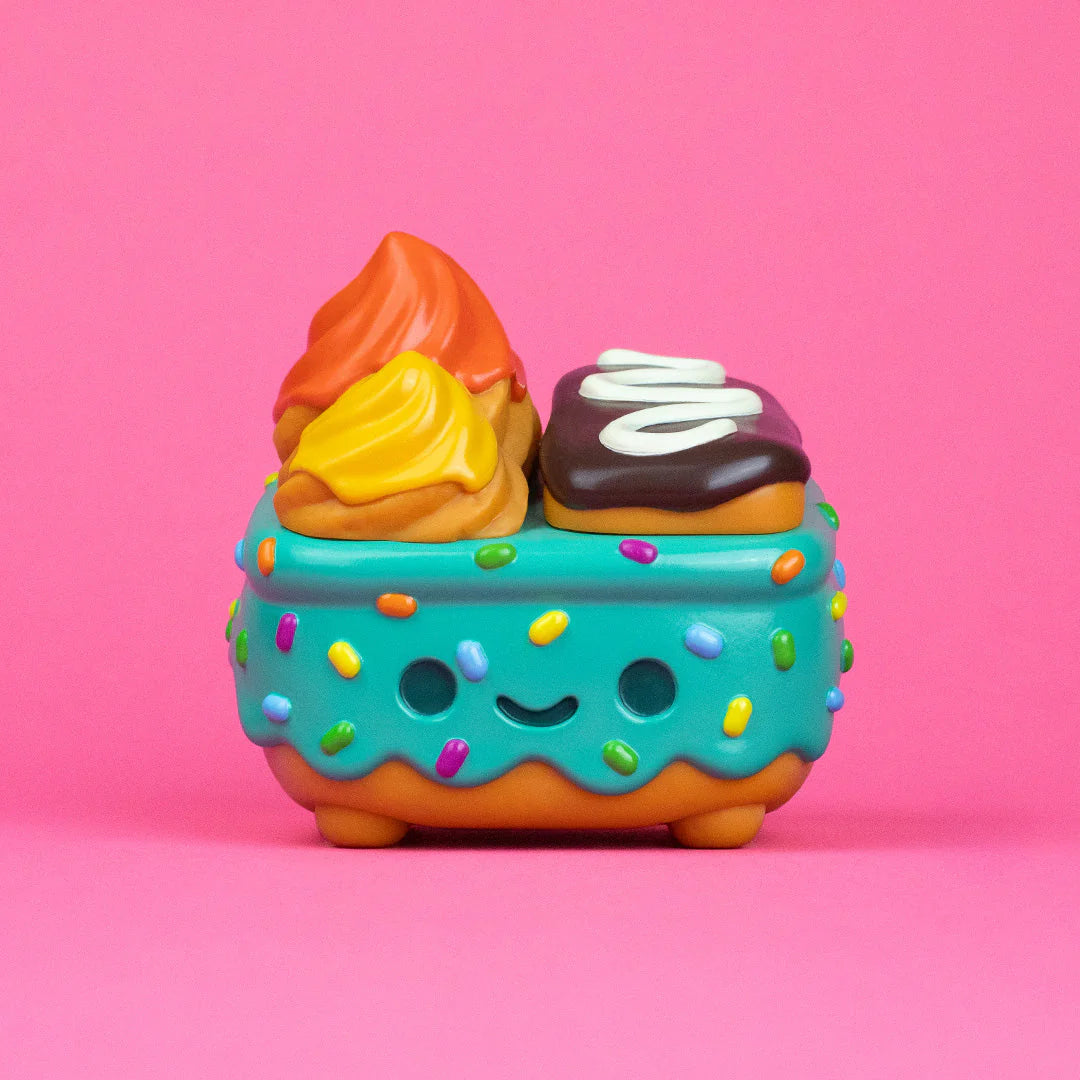A cute, cartoon-style dessert toy from 100% Soft's Dumpster Fire collection, featuring a blue, sprinkle-covered base with a smiling face and swirled, colorful toppings, set against a pink background.