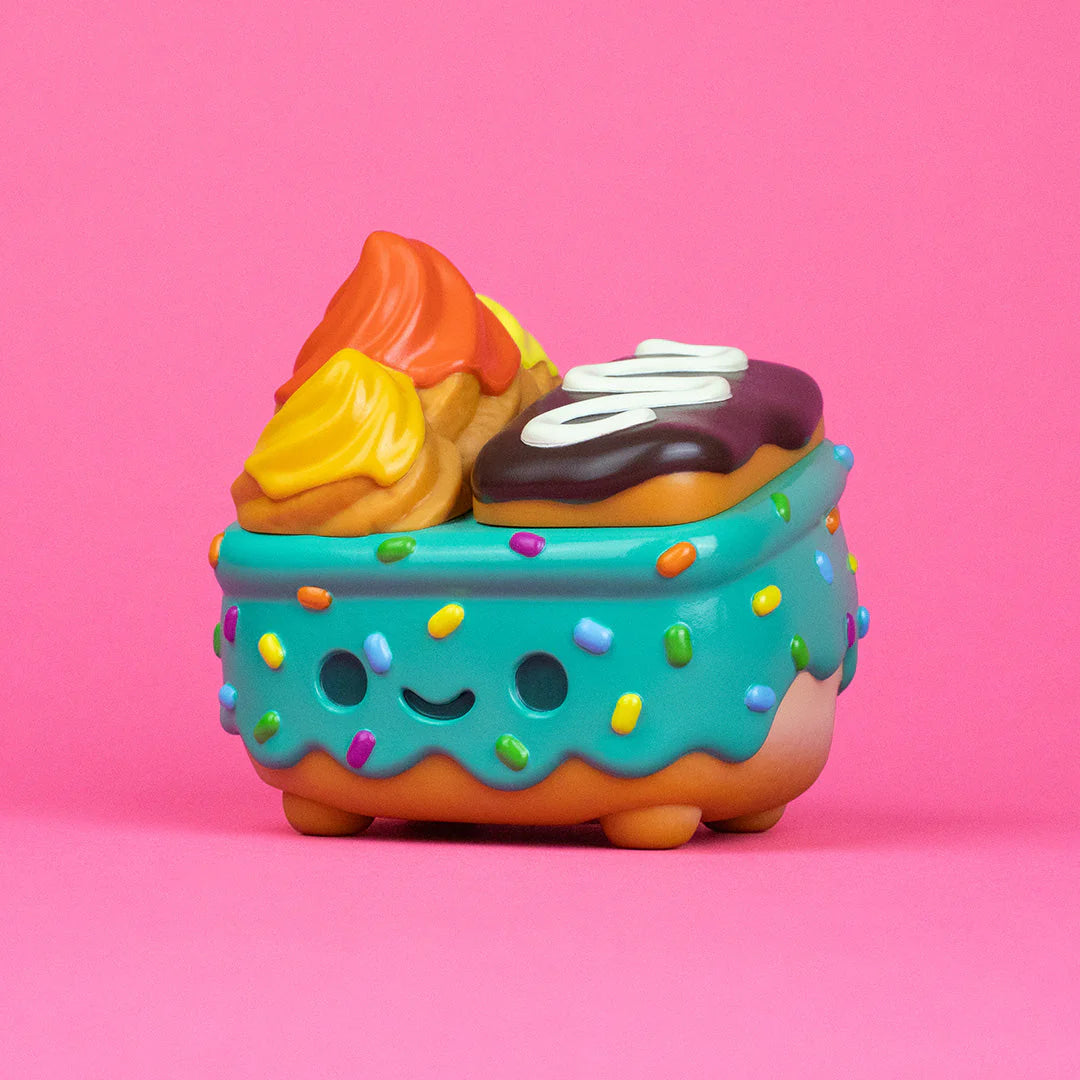 A whimsical dessert character from the 100% Soft Dumpster Fire collection, this figure features a turquoise frosting base adorned with sprinkles, a cheerful smile, and colorful pastry accents on top—evoking the playful spirit of the Donut Green vinyl figure—all presented against a vivid pink background.