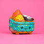 A cute, colorful toy from 100% Soft, shaped like a dessert with blue frosting, sprinkles, a smiling face, and topped with orange and yellow swirls plus a mini chocolate treat—part of the playful Dumpster Fire collection—rests against a pink background.