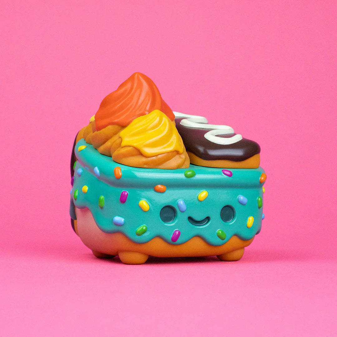 A cute, colorful toy from 100% Soft, shaped like a dessert with blue frosting, sprinkles, a smiling face, and topped with orange and yellow swirls plus a mini chocolate treat—part of the playful Dumpster Fire collection—rests against a pink background.