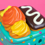 A close-up of a colorful toy from the 100% Soft brand designed to look like frosted pastries and a chocolate treat with white icing, displayed on a teal, sprinkle-covered surface against a pink background—part of the playful Dumpster Fire collection and showcasing the whimsical Donut Green vinyl figure.