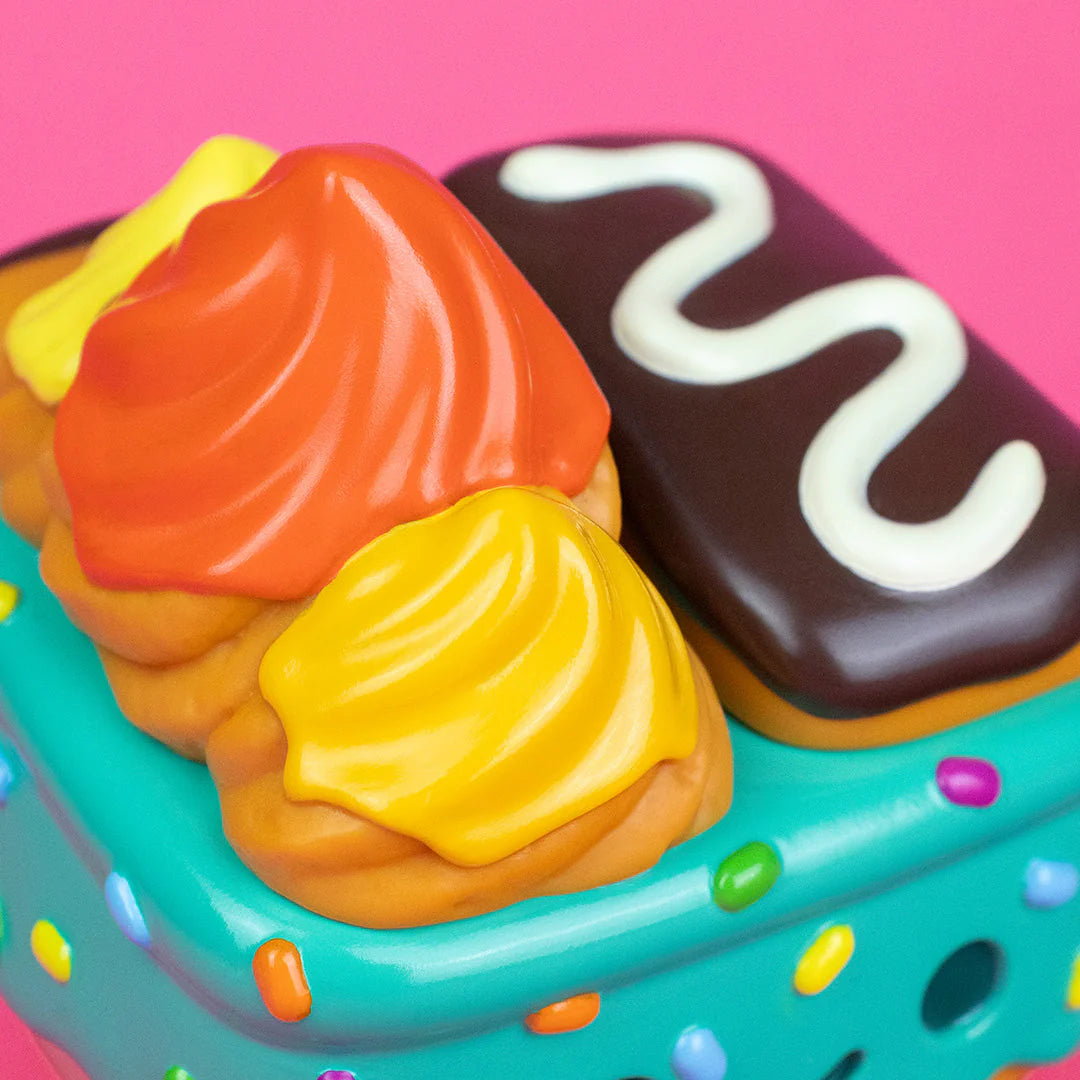 A close-up of a colorful toy from the 100% Soft brand designed to look like frosted pastries and a chocolate treat with white icing, displayed on a teal, sprinkle-covered surface against a pink background—part of the playful Dumpster Fire collection and showcasing the whimsical Donut Green vinyl figure.