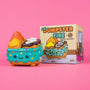 A whimsical collectible from the 100% Soft Dumpster Fire collection, shaped like a decorated ice cream sundae in a blue dumpster with a smiling face and colorful sprinkles, sits beside its matching box against a pink background. This adorable toy captures all the charm that fans of 100% Soft love.