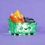 The 100% Soft Gem Dumpster Fire is a small, translucent green and black vinyl figure with a smiling face, four little legs, and orange-yellow crystal flames on its back against a purple background.