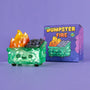 A translucent green vinyl figure from 100% Soft, featuring orange-yellow crystal flames and a smiling face, sits by its colorful "Gem Dumpster Fire" box on a purple surface.