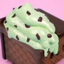 A close-up of mint chocolate chip ice cream with chocolate chunks on a chocolate waffle cone, set against a pink background—evoking the whimsical Mint Chip Vinyl Figure from 100% Soft.