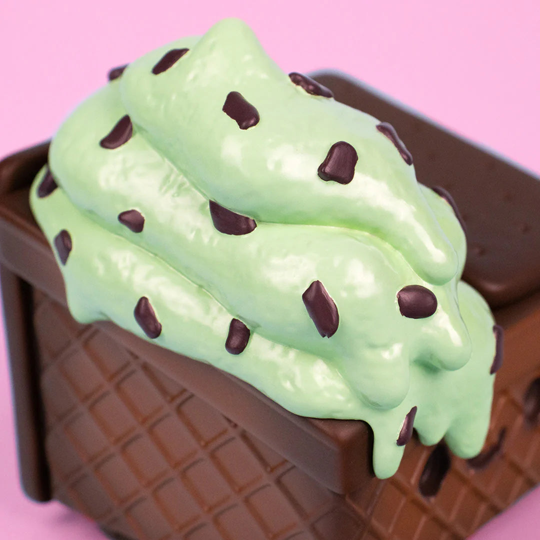 A close-up of mint chocolate chip ice cream with chocolate chunks on a chocolate waffle cone, set against a pink background—evoking the whimsical Mint Chip Vinyl Figure from 100% Soft.