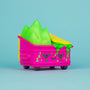 A 100% Soft bright pink vinyl Dumpster Fire figure featuring a smiling face and overflowing with toxic green slime and yellow trash, displayed against a plain light blue background.