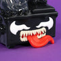 A close-up of a black cartoonish figure with sharp white teeth, a long red tongue sticking out, and white, angry-looking eyes on a purple background—this striking character evokes the 100% Soft Venom Dumpster Fire symbiote collectible, making it a must-have piece for any Marvel vinyl figure enthusiast.