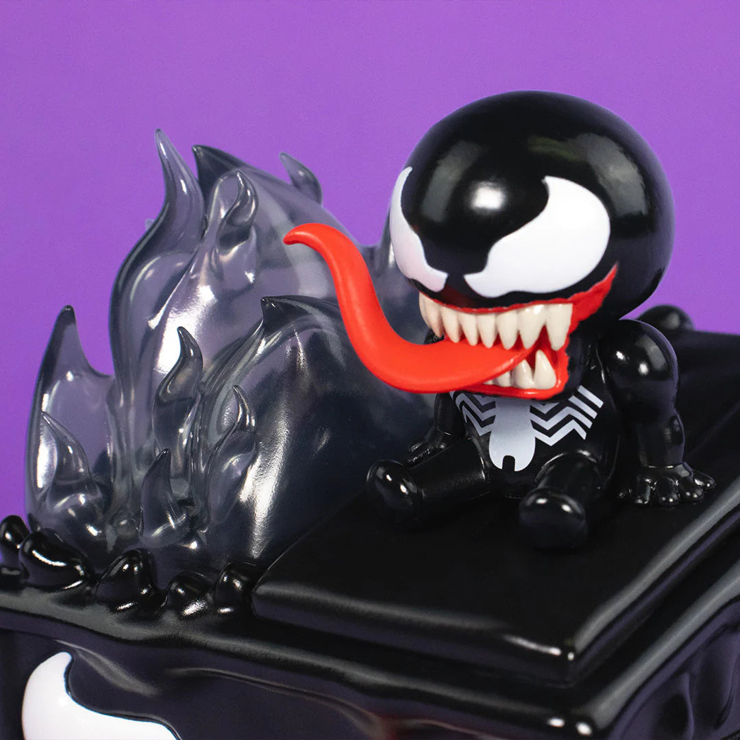 A chibi-style Venom Dumpster Fire figure from 100% Soft, featuring a long red tongue, sits beside translucent black flames on a glossy black base, set against a solid purple background.