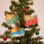 Three colorful Dumpster Fire ornaments from 100% Soft with smiling faces hang on a tree amid festive baubles. This deluxe set features blue, green, and red enamel ornaments, each topped with bright orange flames.