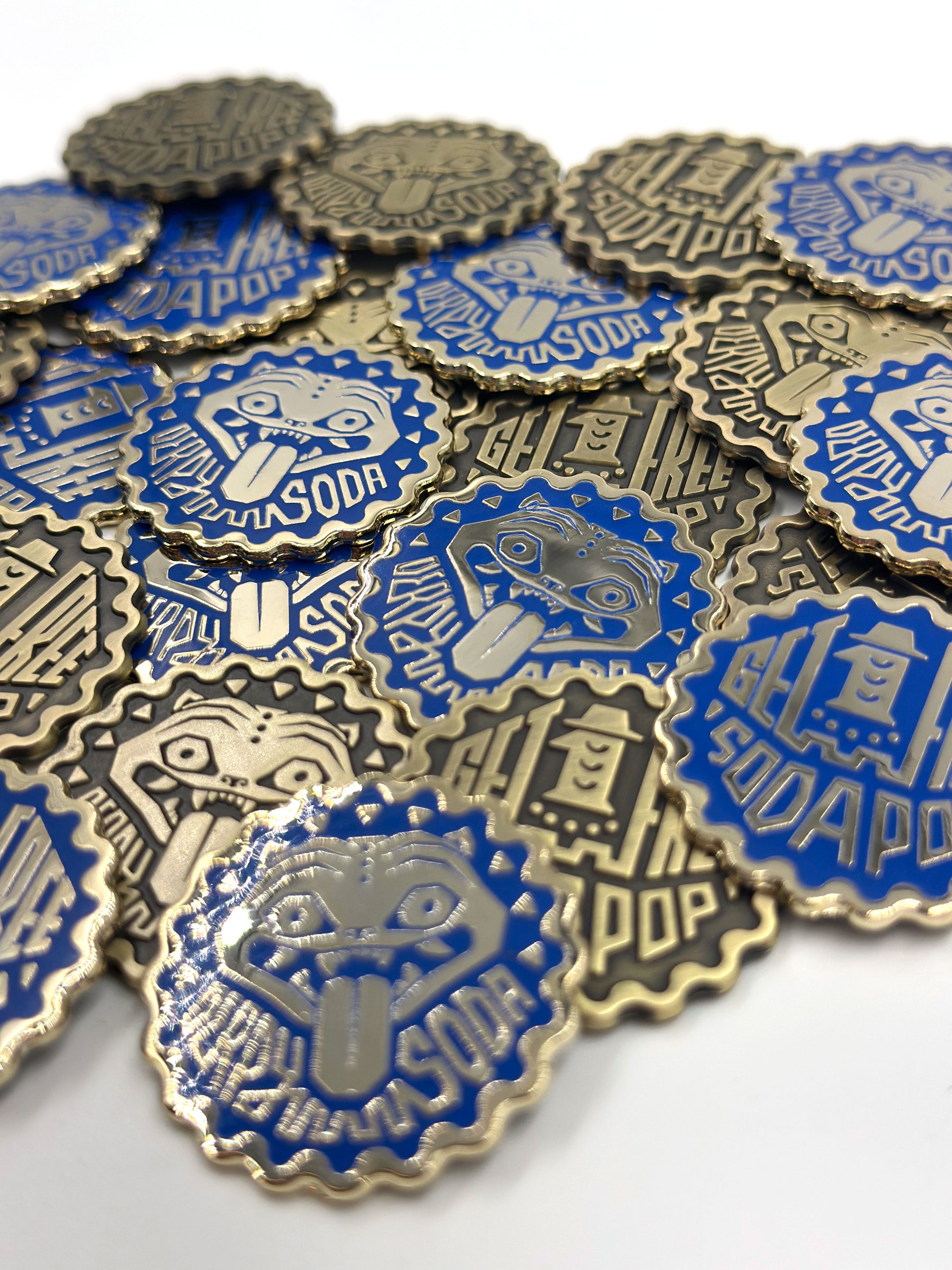 A set of metallic bottle cap-shaped pins by Bruce Yan, featuring blue and silver designs with a cartoon tongue-out face and "GET A FREE SODAPOP" text—ideal as quirky, limited edition collectible metal tokens.