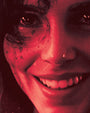A close-up of a smiling person’s face in red and black tones, with one eye and cheek partly covered in dark, splattered markings, giving a dramatic, intense effect; the image evokes an air of obsession reminiscent of Stephen Campanella’s signature archival pigment prints.