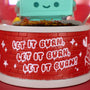 A close-up of a red brick-patterned base with white graffiti-style “LET IT BURN” repeated three times. The bottom of a green, smiling cartoon figure from the 100% Soft Dumpster Fire Snow Globe is visible at the top.