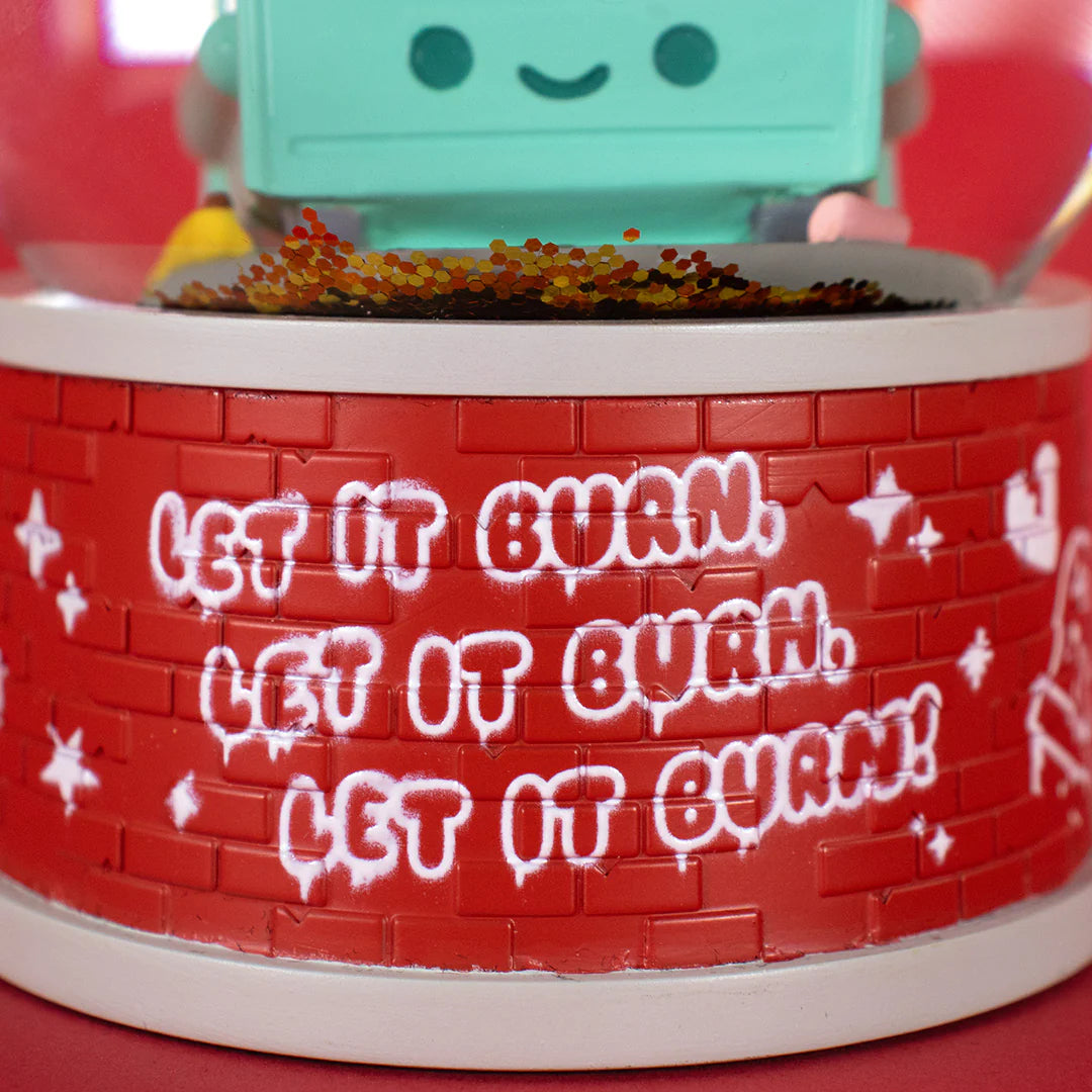 A close-up of a red brick-patterned base with white graffiti-style “LET IT BURN” repeated three times. The bottom of a green, smiling cartoon figure from the 100% Soft Dumpster Fire Snow Globe is visible at the top.