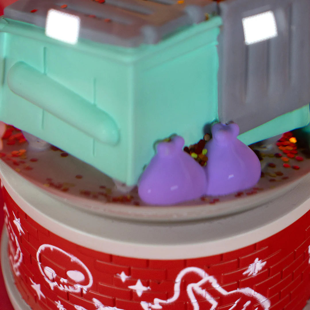 A cake featuring a green and gray dumpster, two small purple trash bags, and red frosting with white drawings—including a skeleton—captures the playful vibe of 100% Soft’s Dumpster Fire Snow Globe.