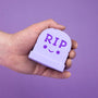 A hand holds a light purple gravestone-shaped stress squishy made from soft polyurethane foam by 100% Soft, featuring a whimsical RIP face on a matching purple background.