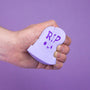 A hand holds a stress squishy resembling a small gravestone with RIP in dark purple, made from polyurethane foam. Designed by 100% Soft, it stands out elegantly against a solid purple background.