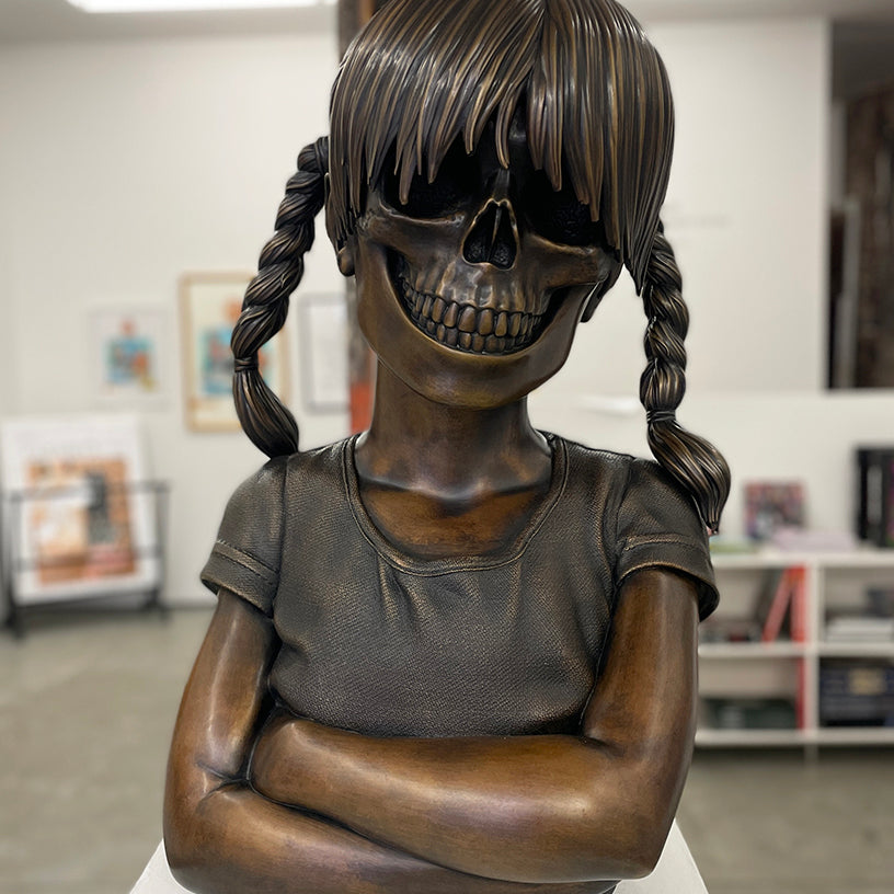 A cast bronze sculpture by Zoltron, titled "Sue Nami Transfiguration," features a girl with braided hair, arms crossed, and a skull in place of her face. Presented by Spoke Art, it's displayed in a modern gallery with framed art and bright lighting.
