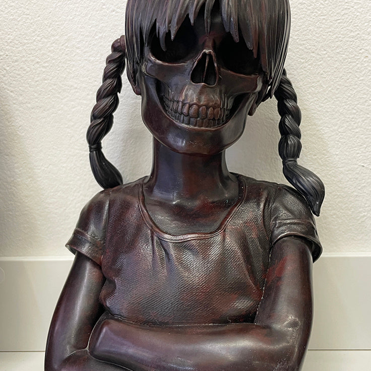 A cast bronze sculpture from Spoke Art by Zoltron, titled Sue Nami: Transfiguration, features a young girl with braided hair, a t-shirt, crossed arms, and a skull face set against a plain light background.