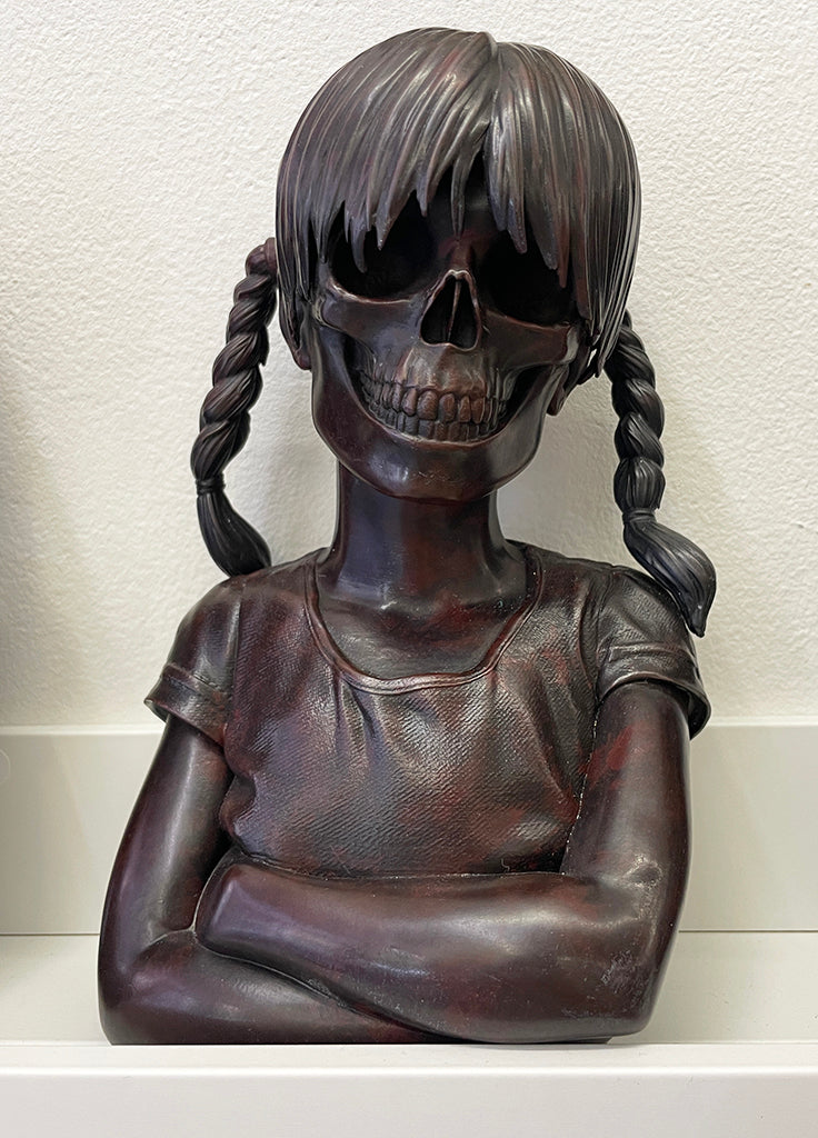 A cast bronze sculpture from Spoke Art by Zoltron, titled Sue Nami: Transfiguration, features a young girl with braided hair, a t-shirt, crossed arms, and a skull face set against a plain light background.