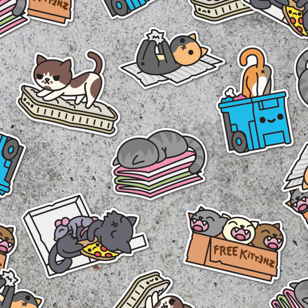 Playful cartoon cat vinyl stickers from 100% Soft feature adorable kitties sleeping on blankets, eating pizza from a box, playing in a recycling bin, and sitting in a Free Kittens box—all scattered across a textured gray background. These weatherproof, die-cut stickers are ready to add some fun and personality to your space!.