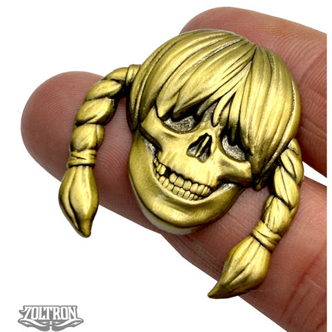 A brass-colored skull pin by ZOLTRON with a wide grin, braided pigtails, bangs, and the VOLTRON logo in the bottom left—this limited edition piece is true Zoltron art.